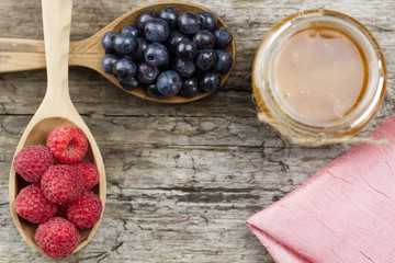 Raspberries and blueberries in spoons with pink napkin and a jar of honey on wooden background. Healthy eating, diet