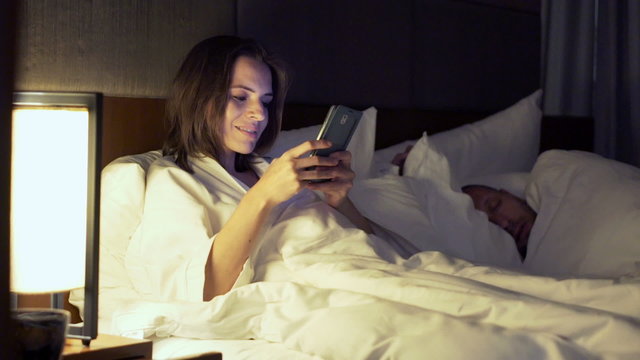 Couple in bed at night, woman texting with lover on smartphone, her husband sleeping
