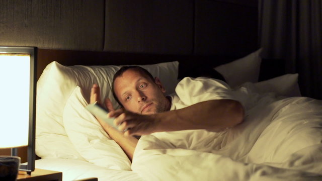 Couple in bed at night, man texting his lover on smartphone, his wife sleeping