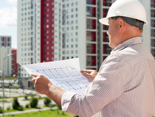 An engineer in white helmet examining blueprints on a background with buildings and green park area