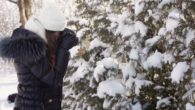Beautiful young woman in winter clothing and knitted white hat photographing snowy forest
