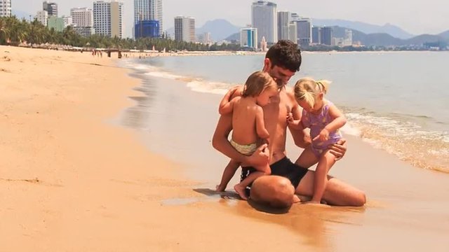 father plays two little girls sitting on edge of sea on beach 