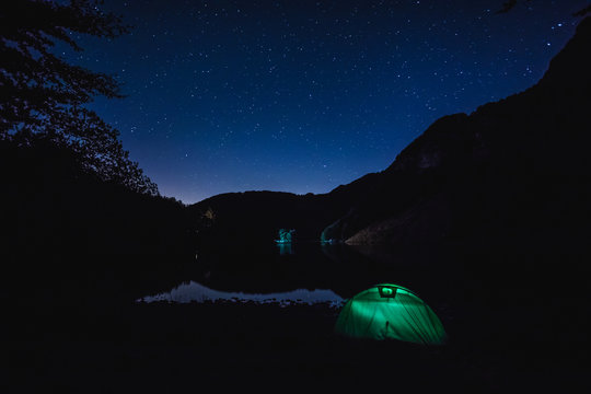 A tent lit, in a summer night under a starry sky is placed in a lake near the italian mountains