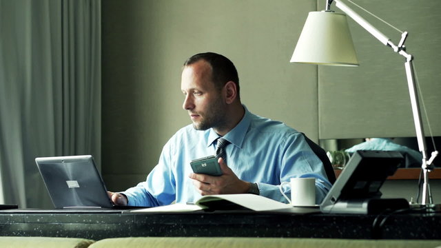 Young businessman comparing data on smartphone, laptop and documents sitting in office
