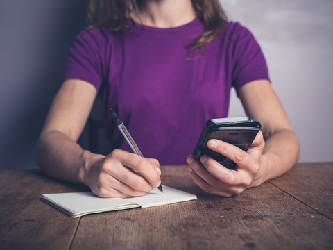 Young woman using phone and writing in notepad