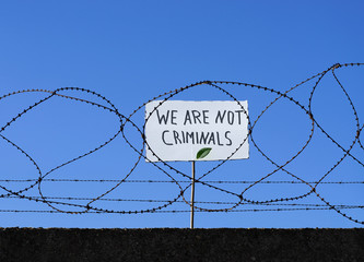 Jemand hinter Stacheldraht hält Schild mit Aufschrift WE ARE NOT CRIMINALS hoch