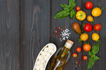 Cherry tomatoes of various color, mozzarella, basil leaves, spices and olive oil from above over dark wooden table. Italian caprese salad recipe ingredients. Top view, free text copy space