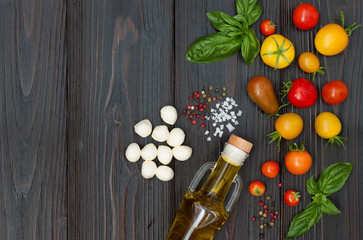 Cherry tomatoes of various color, mozzarella, basil leaves, spices and olive oil from above over dark wooden table. Italian caprese salad recipe ingredients. Top view, free text copy space