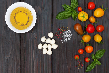Cherry tomatoes of various color, mozzarella, basil leaves, spices and olive oil from above over dark wooden table. Italian caprese salad recipe ingredients. Top view, free text copy space