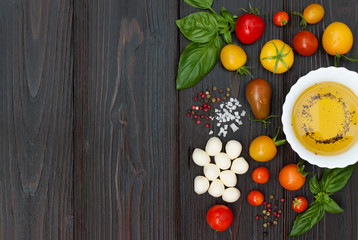 Cherry tomatoes of various color, mozzarella, basil leaves, spices and olive oil from above over dark wooden table. Italian caprese salad recipe ingredients. Top view, free text copy space