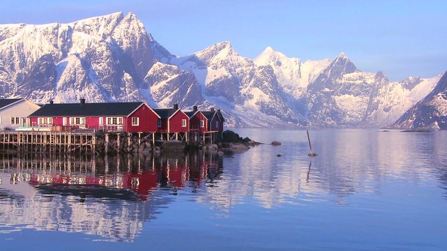 Snowcapped peaks loom behind a red fishing village in the Arctic Lofoten Islands, Norway.
