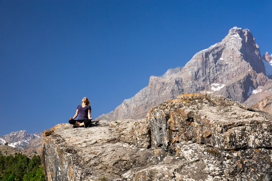 Young woman doing exercise Female Rock Climber Sitting in Yoga Zen Mediation Pose to Stretch Body Mountain Panoramic Landscape Outdoor Sunny Sky Peaks