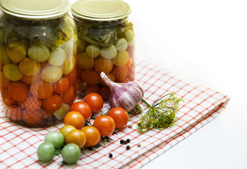 Jars with preserved pickled tomatoes cherry on table with garlic and black pepper isolated on white.  Different degrees of ripeness of tomatoes cherry.