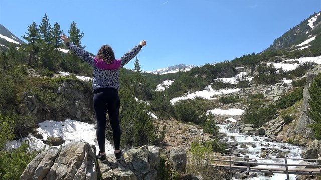 Young girl hiker do victory pose with hands open near mountain peak with snow melting waters spring flow. UHD 4K steadycam stock footage