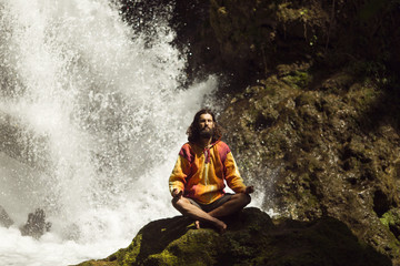 Young man meditating next to waterfall in the mountains - obrazy, fototapety, plakaty