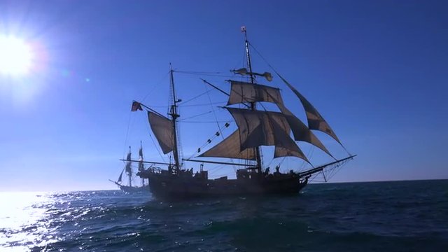 A tall masted clipper ship sails on the high seas against the sun.
