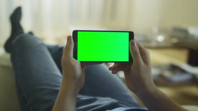 Man is Laying on Couch at Home and Holding Android Phone with Green Screen in Landscape Mode.