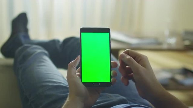 Man is Laying on Couch at Home and Using Android Phone with Green Screen in Portrait Mode on Lap.