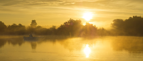 Wędkarz na łodzi łowiący w mglisty poranek nad rzeką - obrazy, fototapety, plakaty