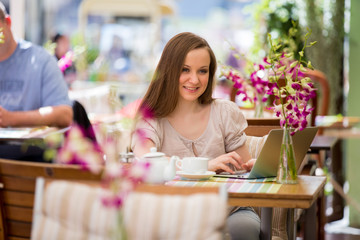 Young beautiful woman sitting in cafe and drinking coffee. Working with the laptop on terrace.

