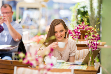 Young beautiful woman sitting in cafe and drinking coffee. Working with the laptop on terrace.
