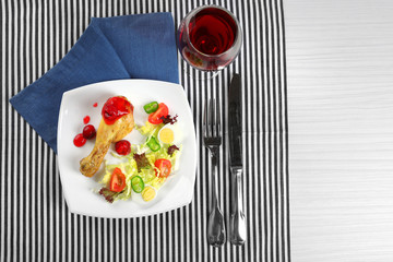 Dish of baked chicken leg and vegetable salad in white plate with glass of wine on table with napkin, top view