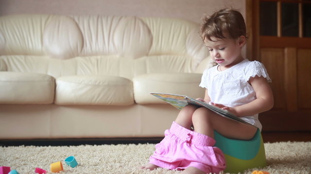 Clever toddler girl sitting on a potty and reading book.
