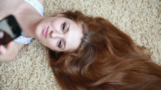 Beautiful girl with red hair lying on the carpet taking selfie. Dolly shot.