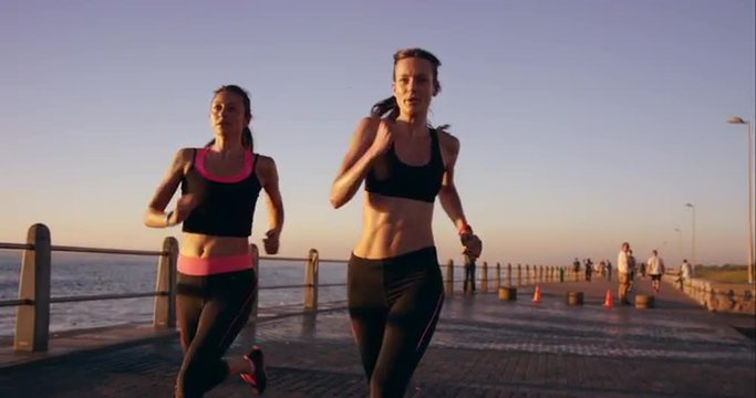 Two athletic woman running outdoors slow motion on promenade at sunset near ocean enjoying evening run