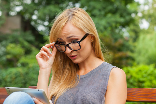 Sensual blonde woman sitting on wooden bench in park. She is using tablet pc.