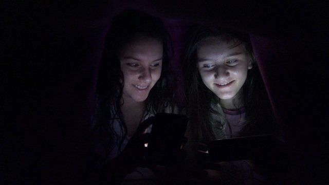 4K happy teen friends surf internet on smartphne, tablet hidden under bed blanket look pictures on social network. UHD stock video, wide shot