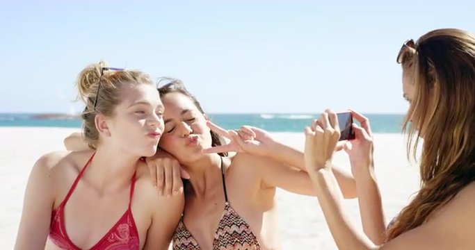 Three teenage girl friends taking photo on beach wearing colorful bikini sharing vacation photo