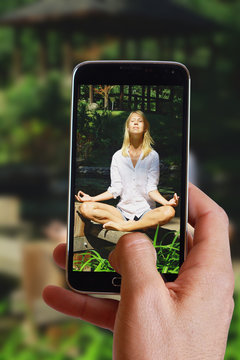 Male hand taking photo of Young Woman meditating in lotus position in japanese garden with cell, mobile phone. Attractive blonde female practices yoga outdoors. Healthy lifestyle concept