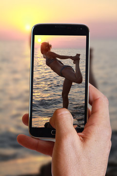 Male hand taking photo of woman meditating on the beach near the sea, ocean, during sunset with cell, mobile phone.