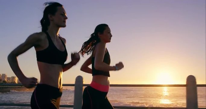 Two athletic woman running outdoors slow motion on promenade at sunset near ocean enjoying evening run