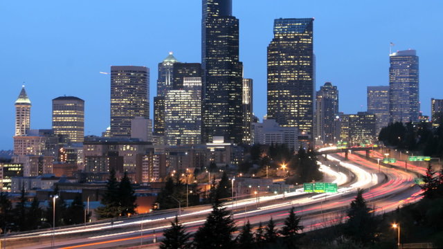 As the golden-hour darkens into night, accelerated traffic blurs into streaks of light before an illuminated Seattle skyline.