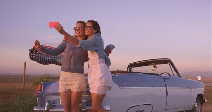 beautiful Girl friends taking selfies on road trip at sunset with vintage car