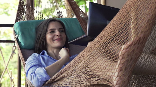 Young woman chatting on laptop while lying on hammock on terrace