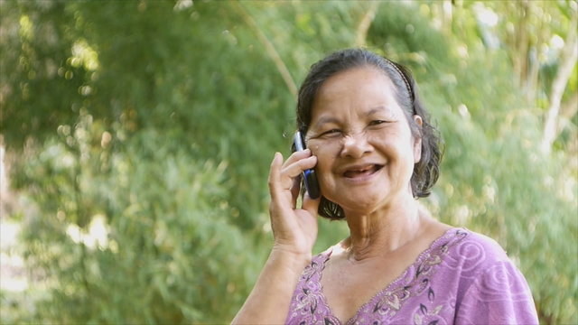 60 Years Old Thai Woman Smiling While Talking On Mobile Phone With Bokeh Green Background