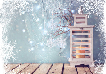 white wooden vintage lantern with burning candle and tree branches on wooden table. retro filtered image with glitter and snowflake overlay
