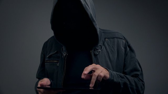 Unrecognizable hooded person with digital tablet computer going on line, using internet and modern gadget to commit cybercrime.
