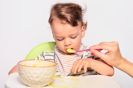 Baby Eating Food With A Spoon, Toddler Eating Messy And Getting Dirty, Infant Having Oatmeal As Breakfast, Parent Hand Feeding His Child