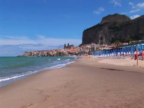 Umbrellas rest on a beach near houses and waves breaking in Cefalu, Italy.  