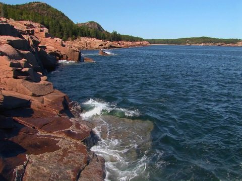 Water crashes against rocks that are near trees and a mountain range in Acadia National Park in Maine.