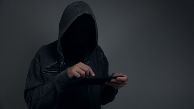 Unrecognizable hooded person with digital tablet computer going on line, using internet and modern gadget to commit cybercrime.