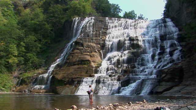 A fisherman stands at the bottom of a wide waterfall waiting for a bite in Ithaca Falls, New York.