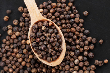 Closeup black pepper whole in wooden spoon on black stone table, top view