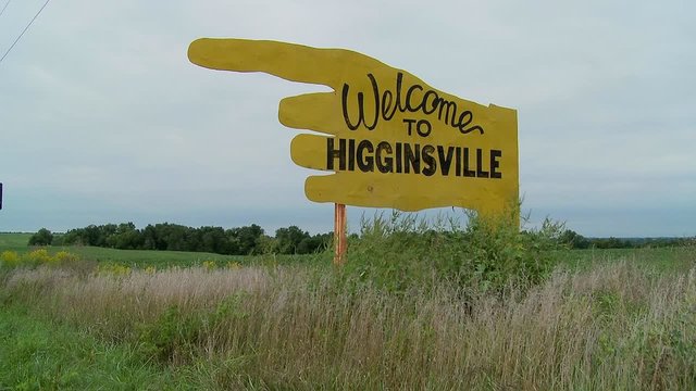 A large yellow sign points to Higginsville, Missouri from a farm field.