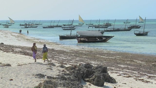 Two Muslim women walk along a beach in Zanzibar with dhow sailboats in the background.