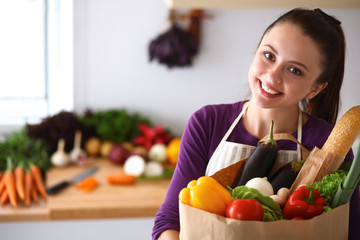 A young woman standing in her kitchen holding a bag of groceries